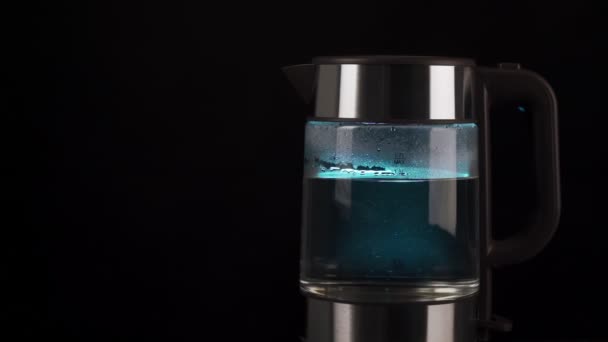 Água fervente em uma chaleira elétrica de vidro sobe em bolhas em câmera lenta. Com retroiluminação azul sobre um fundo preto. — Vídeo de Stock