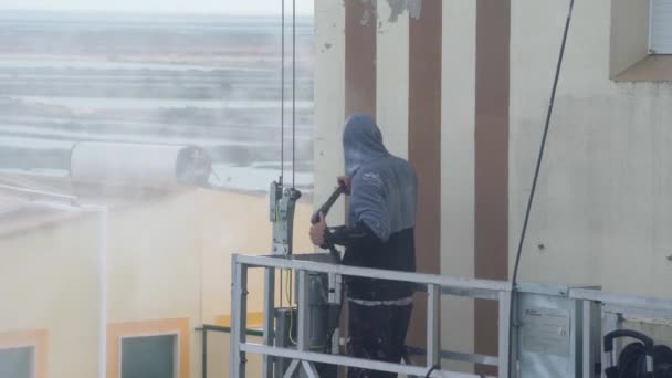 A man uses a high-pressure machine to disinfect the walls of a multi-storey building for subsequent repairs. On an industrial paint hoist. — Vídeo de Stock