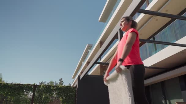 A young woman on the balcony is cleaning the house, shaking off the dust from the rug from the outside. Slow motion video — Wideo stockowe