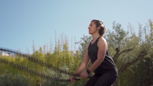 Joven mujer de fitness haciendo ejercicio con dos cuerdas cogidas de la mano moviéndose rápido, al aire libre en el parque. Vídeo en cámara lenta — Vídeos de Stock