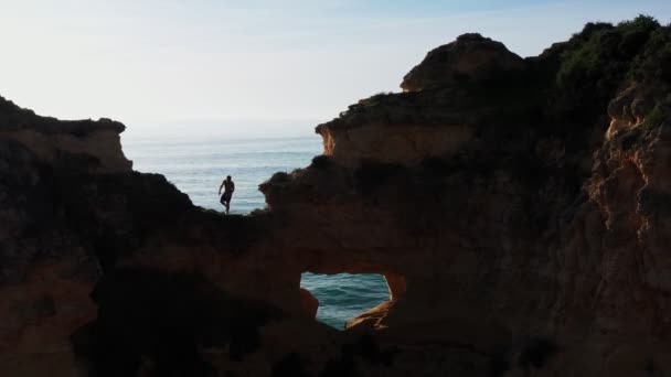 Güneşe karşı siluet dağ yamacında genç bir adam denize doğru aşırı bir sıçrama yapmak üzere.. — Stok video