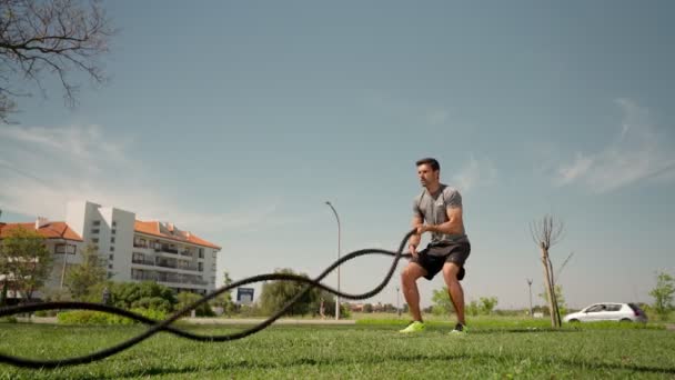 Мускулистый кавказский спортсмен создает волны с боевой веревкой на траве снаружи. Профессиональный спортсмен занимается верёвками на открытом воздухе. — стоковое видео
