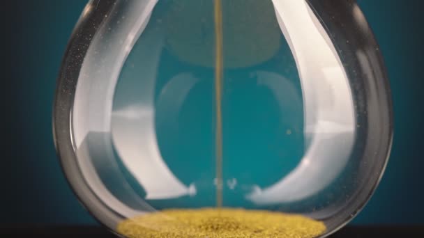 Un reloj de arena hecho de virutas de metal amarillo pasa a través de un embudo, simbolizando el concepto de tiempo en movimiento. — Vídeos de Stock