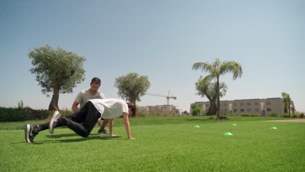 一个有个人健身教练的年轻人进行俯卧撑和抵抗，以求得耐力和身体的协调。在有棕榈树的草地上，. — 图库视频影像
