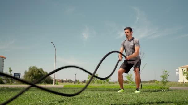 Atleta caucásico muscular hace olas con una cuerda de batalla en la hierba exterior. Un atleta profesional se dedica a las cuerdas al aire libre. mociones lentas — Vídeo de stock