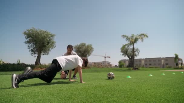 Młody facet z osobistym trenerem fitness wykonuje pompki, a opór biegnie dla wytrzymałości i koordynacji ciała. Na łące z palmami. — Wideo stockowe
