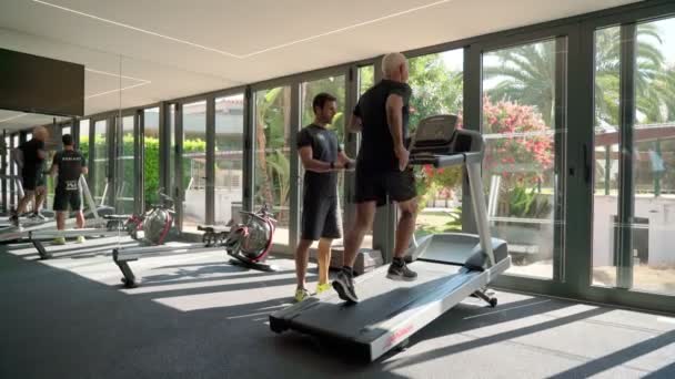Пожилой пенсионер в спортзале бегает на беговой дорожке с помощью личного тренера. Португалия Виламоура 13 марта 2021 года — стоковое видео