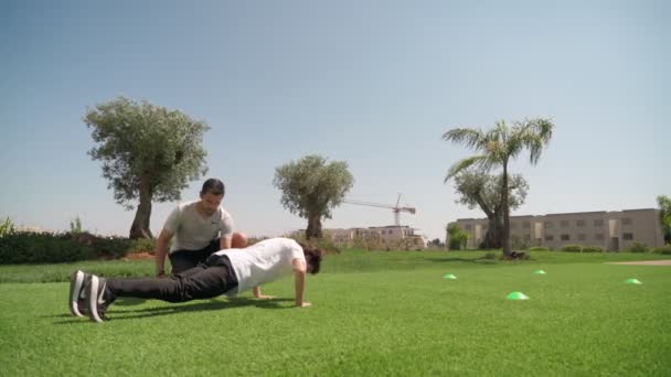 Un joven con un entrenador de fitness personal realiza flexiones y carreras de resistencia para la resistencia y la coordinación del cuerpo. En un prado con palmeras. — Vídeo de stock