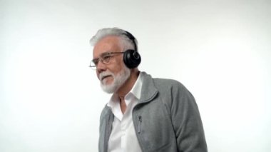 Gri sakallı ve kulaklıklı şık, yaşlı bir adam modern müzik dinliyor ve izole edilmiş beyaz bir arka planda dans ediyor. Yavaş çekim