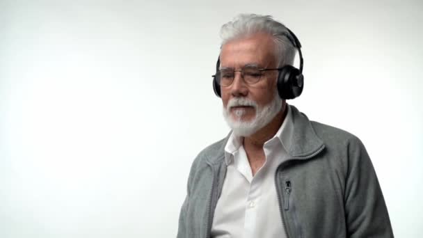 Стильний емоційний старий з сірою бородою і навушниками слухає сучасну музику і танці на білому ізольованому тлі. Повільний рух — стокове відео