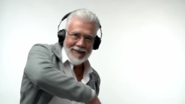 Snygg känslomässig gammal man med grått skägg och hörlurar lyssnar på modern musik och danser på en vit isolerad bakgrund. Långsamma rörelser — Stockvideo