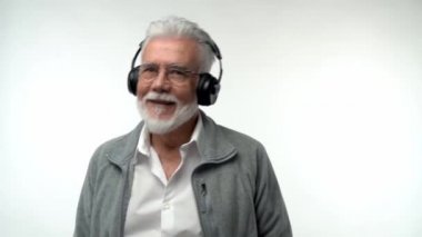 Gri sakallı ve kulaklıklı şık, yaşlı bir adam modern müzik dinliyor ve izole edilmiş beyaz bir arka planda dans ediyor. Yavaş çekim