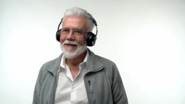 Stylish emotional old man with a gray beard and headphones listens to modern music and dances on a white isolated background. Slow motion — Stock Video
