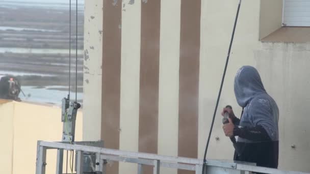 Bir adam çok katlı bir binanın duvarlarını dezenfekte etmek için yüksek basınçlı bir makine kullanıyor. Endüstriyel boya asansöründe. — Stok video