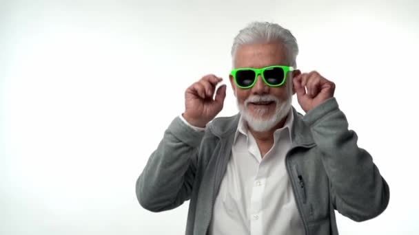 Pensionista de velho emocional elegante com uma barba cinza em óculos verdes dança com emoções engraçadas em um fundo isolado branco. Alegre engraçado despreocupado velhice — Vídeo de Stock