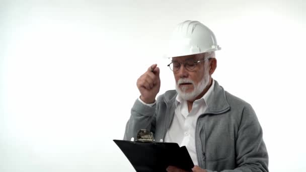 A bearded elderly pensioner man in a construction helmet writes annotations with a pen on a writing board on a white background. — Vídeos de Stock
