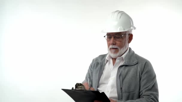 En skäggig äldre pensionär i bygghjälm skriver anteckningar med en penna på skrivtavla på vit bakgrund. — Stockvideo