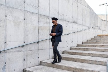 Şık bir şapka ve güneş gözlüğü takan genç Latin eşcinsel adam, beyaz arka planda izole bir şekilde akıllı telefonuyla mesajlaşırken merdivenlerden iniyor. LGTB. Boşluğu kopyala