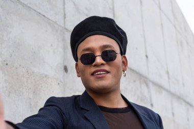 Makyajlı, şık bir şapka takan, güneş gözlüğü takan, beyaz bir arka planda izole edilmiş selfie çeken genç, eşcinsel bir adam. LGBT. 