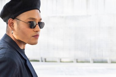 Makyajlı, şık bir şapka ve güneş gözlüğü takan, beyaz bir arka planda kameraya bakan genç, eşcinsel bir adam. LGBT. 