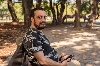 Parktaki bankta oturan bir adam elinde cep telefonuyla kameraya bakıyor.