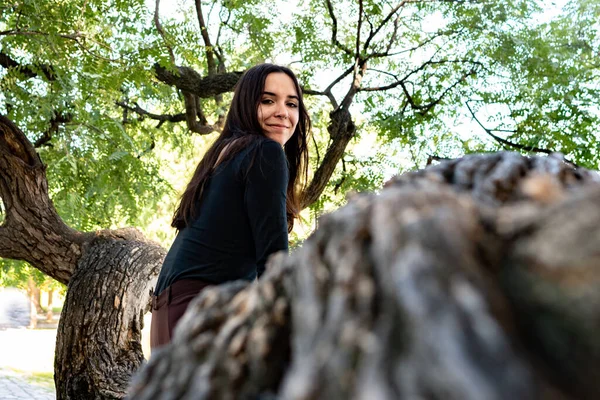 Ağaç dalında oturan bir kadın halka açık bir parkta poz verip kameraya bakıyor.