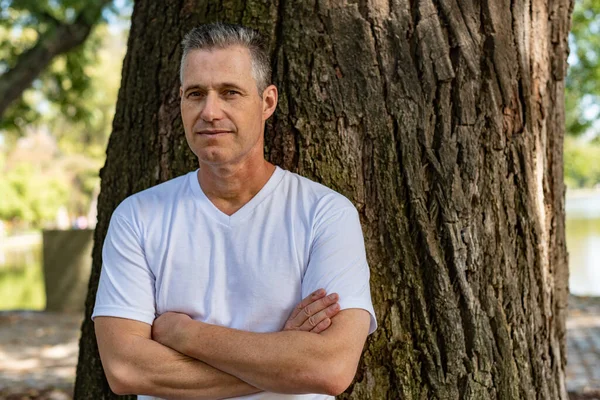 A handsome man leaning on a tree with his arms folded and looking at the camera