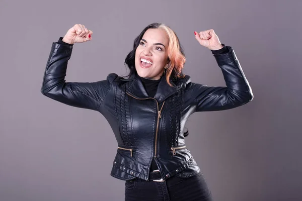 Portrait of a successful woman in a leather jacket while raising hand in success gesture over gray background. Concept of confidence and woman empowerment.