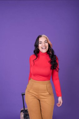 Happy woman traveler holding her suitcase while walking against purple background. Studio shot