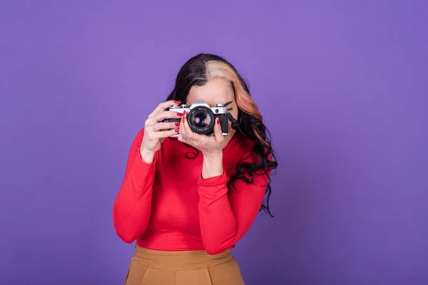 Attractive young lady taking a photo with her film camera over a violet background.  Studio shot.