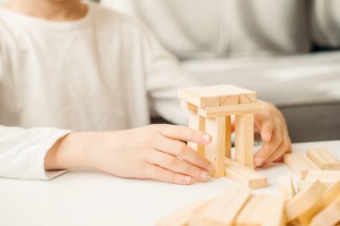 Beyaz tişörtlü, yakın plan bir çocuğun elleri masanın üzerine bir ev inşa ediyor. Çocuklar ve yetişkinler için masa oyunları. Aletsiz bir eğlence. Evde tatil oyunları var. Çocuk tahta küplerle oynuyor..