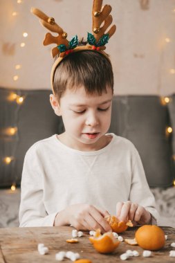 Noel boynuzlu çocuk, oturur ve sarı bir çelengin arka planına mandalina soyar. Evde beyaz tişörtlü bir çocuk, marşmelovlu ahşap bir masada mandalina soyuyor.