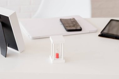 Ofiste beyaz bir masaüstü, kum saatiyle, arka planda bir telefon ve tablet var.