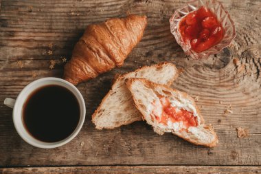 Tereyağlı kruvasan ve çilek reçeli ile bir fincan kahve, ahşap arka planda, üst manzara.