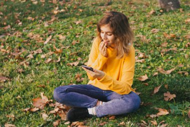Fall mood. Young girl in yellow sweater with smartphone sitting on autumn grass
