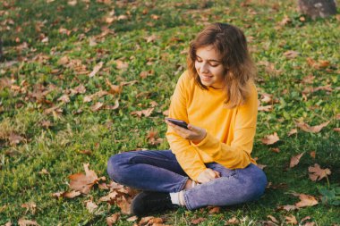 Fall mood. Young girl in yellow sweater with smartphone sitting on autumn grass