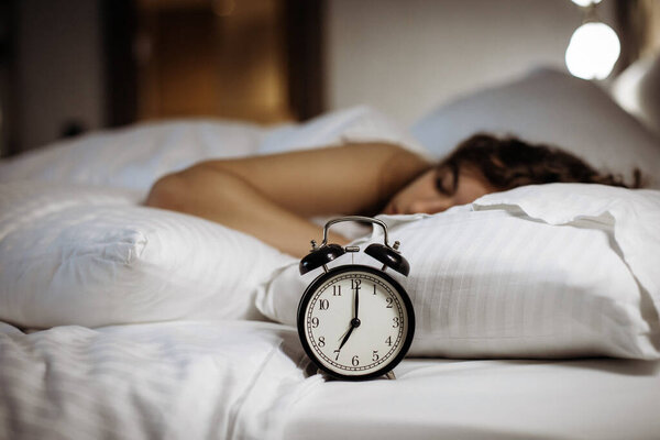 Будильник на кровати со спящей женщиной позади.
