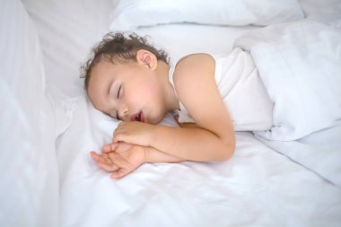Sweet baby boy sleeps on white bed. Curly toddler sleeping