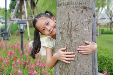 Güzel bir Asyalı kız çocuğu bahçede kollarıyla ağaç gövdesine sarılıyor. Doğanın tadını çıkaran çocuk aşk, doğada gülümseyen.