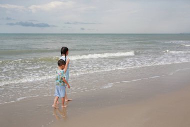 Rear view Asian young sister and little brother walking together on tropical sand beach at sunrise. Happy family boy and girl enjoy in summer holiday.