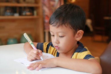Intended Asian little boy drawing in a book with a pen on table in classroom.