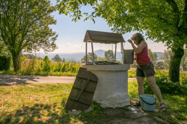 A woman is taking the water from old, stone, water well  with a windlass and crank in the polish village. In the background view of the Cergowa Hill in the Low Beskids, Beskid Niski, Poland