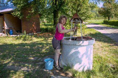 A woman is taking the water from old, stone, water well  with a windlass and crank in the polish village. In the background view of wooden, log house in the Low Beskids, Beskid Niski, Poland