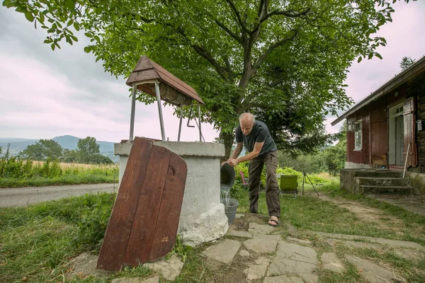 A man is taking the water from old, stone, water well  with a windlass and crank in the polish village. In the background view of the Cergowa Hill in the Low Beskids, Beskid Niski, Poland