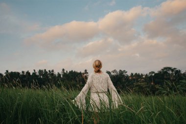 Young woman in flown white dress walking through tall grass field