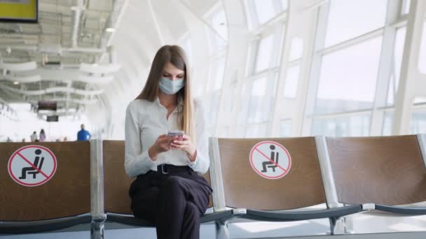 Hermosa mujer de negocios en una máscara en el aeropuerto utilizando el teléfono envía mensajes de texto, mira a la cámara, comprobar los correos electrónicos en Internet, una mujer exitosa gerente en el trabajo. — Vídeos de Stock