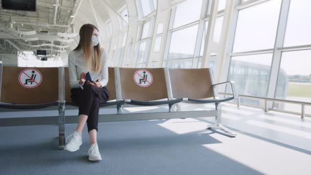 Donna d'affari sicura con una maschera seduta all'aeroporto, in possesso di un passaporto e biglietti aerei, mantiene una distanza in una pandemia. Ti aspetto all'aeroporto. Concetto di viaggio pandemico. Distanza sociale — Video Stock