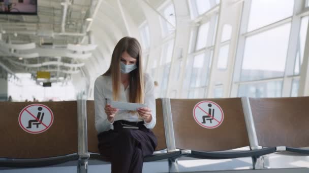 Una mujer de negocios segura con una máscara sentada en el aeropuerto, con pasaporte y billetes de avión, mantiene una distancia en una pandemia. Esperando en el aeropuerto. Concepto de viaje. Mujer mirando entradas — Vídeos de Stock