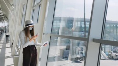 Genç bir kız, modern bir havaalanında elinde biletlerle pencerenin önünde duran bir iş kadını ve bir telefon kameraya bakıyor. Maskeli güzel bir kızın portresi. Uçakla iş gezisi.