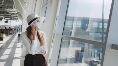 Koruyucu maskeli genç bir kadın havaalanı koridorunda pencerenin yanında duruyor ve akıllı telefon kullanıyor. Havaalanı terminalinin yanında telefonla konuşan küçük bir kızın portresi. İş gezisi konsepti